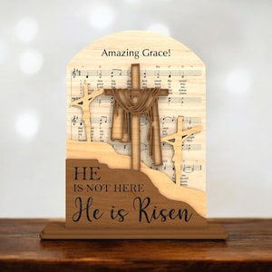 May include: Wooden Easter decoration featuring three crosses, with the central cross draped with fabric. The background includes sheet music and the words "Amazing Grace!" and "He is not here, He is Risen."