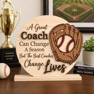 May include: A wooden baseball-themed sign with the text "A Great Coach Can Change A Season But The Best Coaches Change Lives." A baseball glove and ball are featured on the sign. A baseball and a trophy are also visible.
