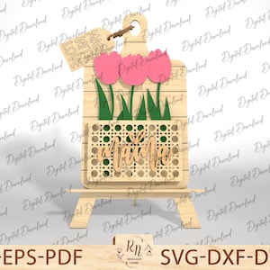 May include: A wooden cutout of a flower pot with the word "mama" in the center. The pot is decorated with a lattice pattern and has three pink flowers with green leaves. A tag with the text "Made with love" is attached to the top of the pot.