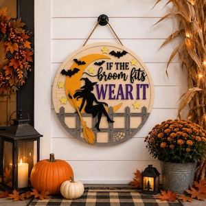 May include: A round wooden sign featuring a black silhouette of a witch sitting on a fence against a yellow crescent moon. The sign includes the text "IF THE broom fits WEAR IT" in black and purple, accented with small yellow stars and black bats.