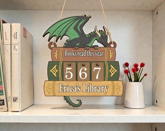 May include: Wooden dragon-themed library sign with the text "Books read this year" and "Erica's Library." The sign has a dragon perched on a stack of books with the numbers 567. A small white vase with red flowers is on the shelf.