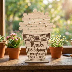 May include: Wooden earring holder in the shape of a flower pot with the text "Thanks for helping me grow." The holder has several holes for earrings and is decorated with floral designs. The holder is on a small wooden base.