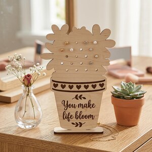 Könnte beinhalten: Ein Ohrringhalter aus Holz in Form eines Blumentopfs mit der Aufschrift "You make life bloom". Der Halter hat mehrere Löcher für Ohrringe und herzförmige Verzierungen. Eine kleine Sukkulente im Topf ist ebenfalls zu sehen.