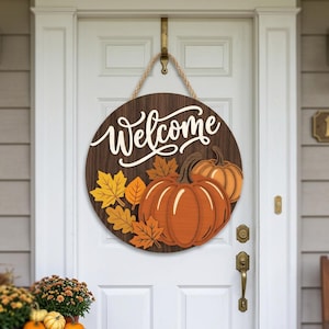 Peut inclure: Une plaque de porte ronde en bois avec une finition grain de bois brun foncé portant le mot Welcome en lettres cursives blanches. La plaque est décorée de deux citrouilles orange et de plusieurs feuilles d'automne jaunes et orange.