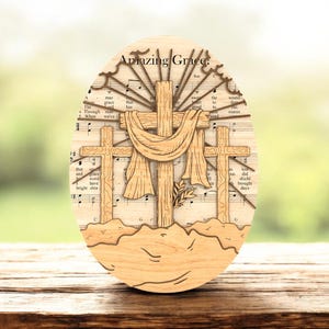 May include: Wooden Easter egg decoration featuring three crosses, a draped cloth, and the words "Amazing Grace." The background is sheet music. The design is made of light-colored wood and stands on a wooden surface.
