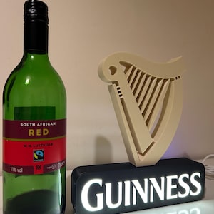 May include: A black and white Guinness sign with a white harp logo and the word "GUINNESS" in white letters. The sign is lit up with white lights. A bottle of South African red wine is to the left of the sign.