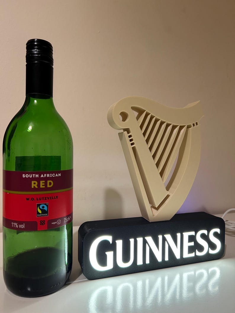 May include: A black and white Guinness sign with a white harp logo and the word "GUINNESS" in white letters. The sign is lit up with white lights. A bottle of South African red wine is to the left of the sign.