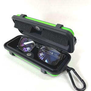 Könnte beinhalten: Eine Brille mit schwarzem Rahmen und reflektierenden Gläsern in einem geöffneten, rechteckigen Hartschalenetui. Das Etui ist schwarz mit leuchtend grünen Rändern, verfügt über eine schwarze Schaumstoffpolsterung und einen schwarzen Karabinerhaken.