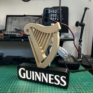 May include: A 3D printed Guinness harp logo with a black base and white LED lights. The logo is illuminated and the word "GUINNESS" is visible on the base.