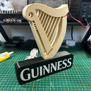 May include: A 3D printed Guinness sign with a black base and a white inscription. The sign features a beige harp with a white outline.