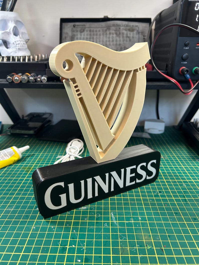 May include: A 3D printed Guinness sign with a black base and a white inscription. The sign features a beige harp with a white outline.