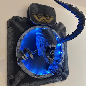 May include: A 3D printed wall mount of an Alien Xenomorph head with a blue light behind it. The Xenomorph is emerging from a circular opening with a Weyland-Yutan Corp. logo above it.