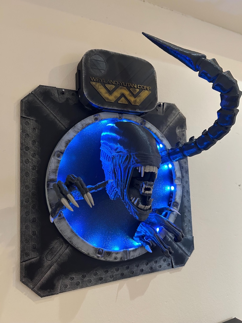 May include: A 3D printed wall mount of an Alien Xenomorph head with a blue light behind it. The Xenomorph is emerging from a circular opening with a Weyland-Yutan Corp. logo above it.