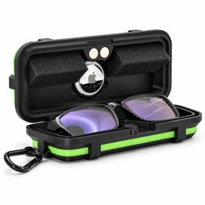 Può includere: Una custodia protettiva rigida nera con una striscia decorativa verde brillante, contenente un paio di occhiali con montatura nera e lenti azzurre, e un Apple AirTag bianco circolare fissato nel coperchio con schiuma nera e un moschettone.