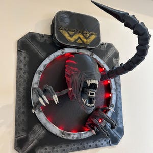 May include: A black and gray 3D printed wall sculpture of an alien from the movie Alien. The alien is depicted with its mouth open, revealing sharp teeth. The sculpture is lit with red LED lights. The sculpture is mounted on a black and gray background with the Weyland-Yutan Corp logo.