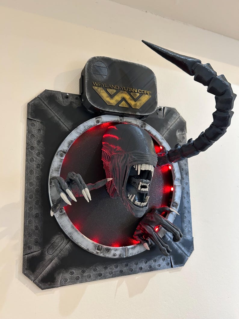 May include: A black and gray 3D printed wall sculpture of an alien from the movie Alien. The alien is depicted with its mouth open, revealing sharp teeth. The sculpture is lit with red LED lights. The sculpture is mounted on a black and gray background with the Weyland-Yutan Corp logo.
