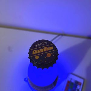 May include: A blue light-up bottle with a black bottle cap that says "Nuka Cola Quantum" in yellow letters. The bottle is sitting on a white surface with a remote control for the light.