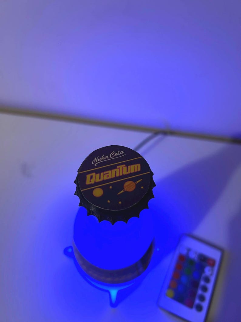 May include: A blue light-up bottle with a black bottle cap that says "Nuka Cola Quantum" in yellow letters. The bottle is sitting on a white surface with a remote control for the light.