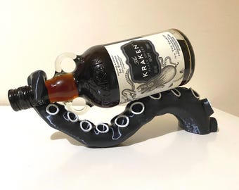 以下が含まれることがあります： 黒いタコの形をしたボトルホルダーに、クラーケンブラックスパイスドラムのボトルが触手で置かれています。