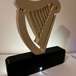 May include: A white 3D printed harp sculpture with a black base and a white cord plugged into the base. The harp is illuminated from behind by a white light.