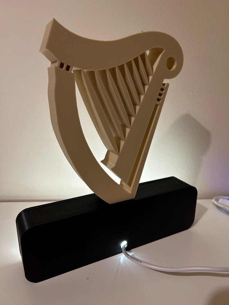 May include: A white 3D printed harp sculpture with a black base and a white cord plugged into the base. The harp is illuminated from behind by a white light.