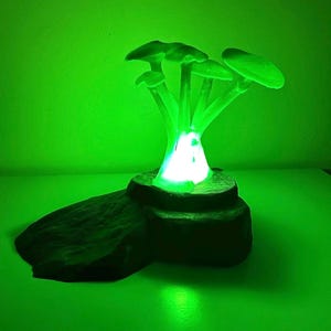 May include: A green mushroom lamp with a bright white light at its core. The lamp features multiple mushroom caps and stems, resting on a dark brown, textured base. The background is a solid green.