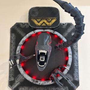 May include: A black and gray 3D printed wall sculpture of an alien head with a red light ring around it. The alien has a large mouth with sharp teeth and a long tail. The sculpture is mounted on a black background with the text "Weyland-Yutani Corp" in gold letters.