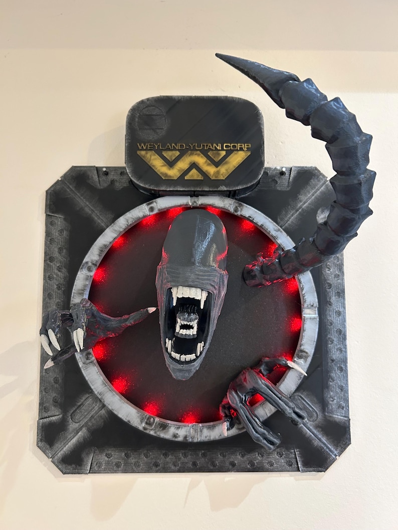 May include: A black and gray 3D printed wall sculpture of an alien head with a red light ring around it. The alien has a large mouth with sharp teeth and a long tail. The sculpture is mounted on a black background with the text "Weyland-Yutani Corp" in gold letters.