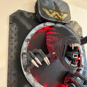 May include: A black and gray 3D printed wall sculpture of an alien creature with glowing red eyes, emerging from a circular opening. The sculpture is mounted on a black and gray background with the text "Weyland-Yutani Corp" on a metal plaque.