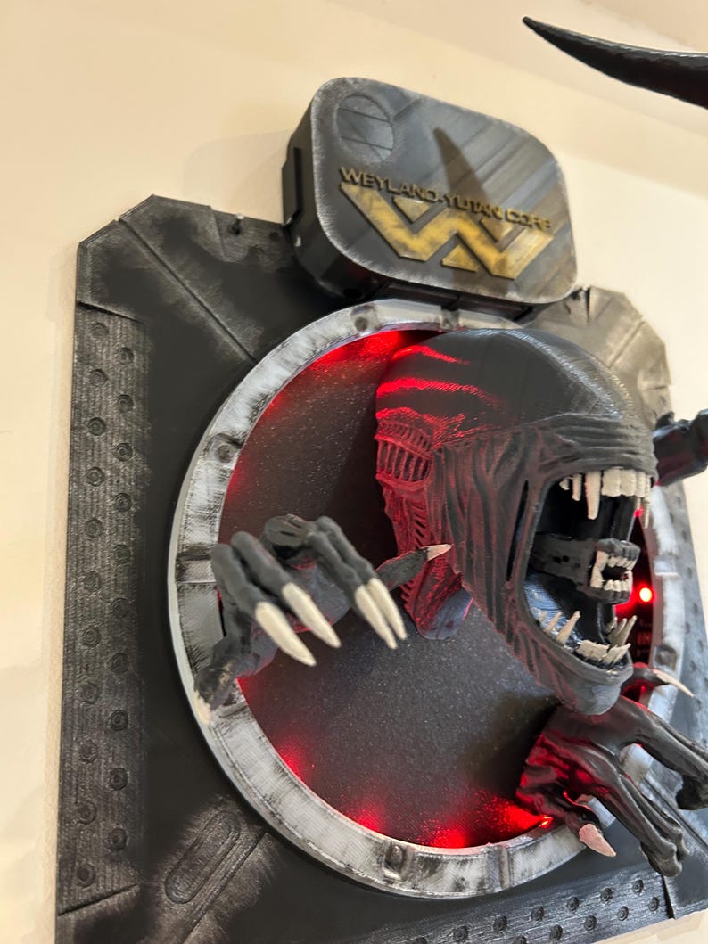 May include: A black and gray 3D printed wall sculpture of an alien creature with glowing red eyes, emerging from a circular opening. The sculpture is mounted on a black and gray background with the text "Weyland-Yutani Corp" on a metal plaque.