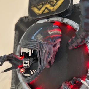 May include: A black and gray 3D printed alien bust with a large mouth and sharp teeth. The bust is mounted on a black and gray wall plaque with the text "Weyland-Yutani Corp" in gold lettering. The plaque has red LED lights around the edge.
