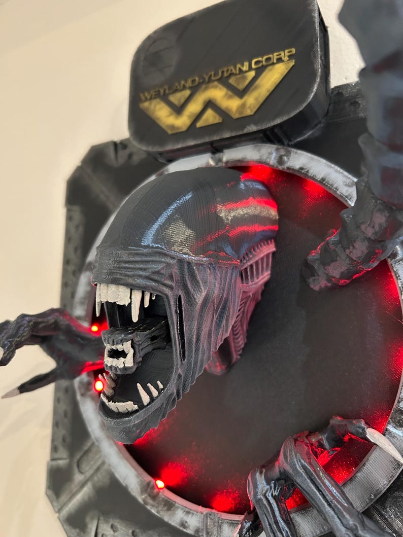May include: A black and gray 3D printed alien bust with a large mouth and sharp teeth. The bust is mounted on a black and gray wall plaque with the text "Weyland-Yutani Corp" in gold lettering. The plaque has red LED lights around the edge.