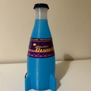 May include: A blue and purple Nuka-Cola Quantum bottle shaped like a rocket ship with a black cord attached. The bottle has a black cap and a white label with the text "Nuka-Cola Quantum" in white and black.