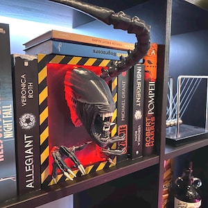 May include: A black Alien creature bookend with a detailed head and claws, emerging from a red-lit space between books. The bookend is placed on a shelf with various book titles visible, including "Night Fall" and "Allegiant".