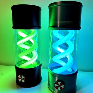 May include: Two cylindrical lamps with black tops and bases, each containing a glowing, spiraling light. One lamp emits green light, the other blue. The bases feature a red and white emblem. The lamps are on a white surface.