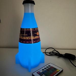 May include: A blue and white rocket-shaped bottle with a label that reads "Nuka Cola Quantum" and a black cap. The bottle is lit up with a blue light and is connected to a remote control with multiple colored buttons.