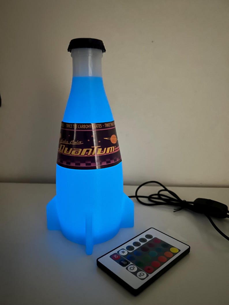 May include: A blue and white rocket-shaped bottle with a label that reads "Nuka Cola Quantum" and a black cap. The bottle is lit up with a blue light and is connected to a remote control with multiple colored buttons.