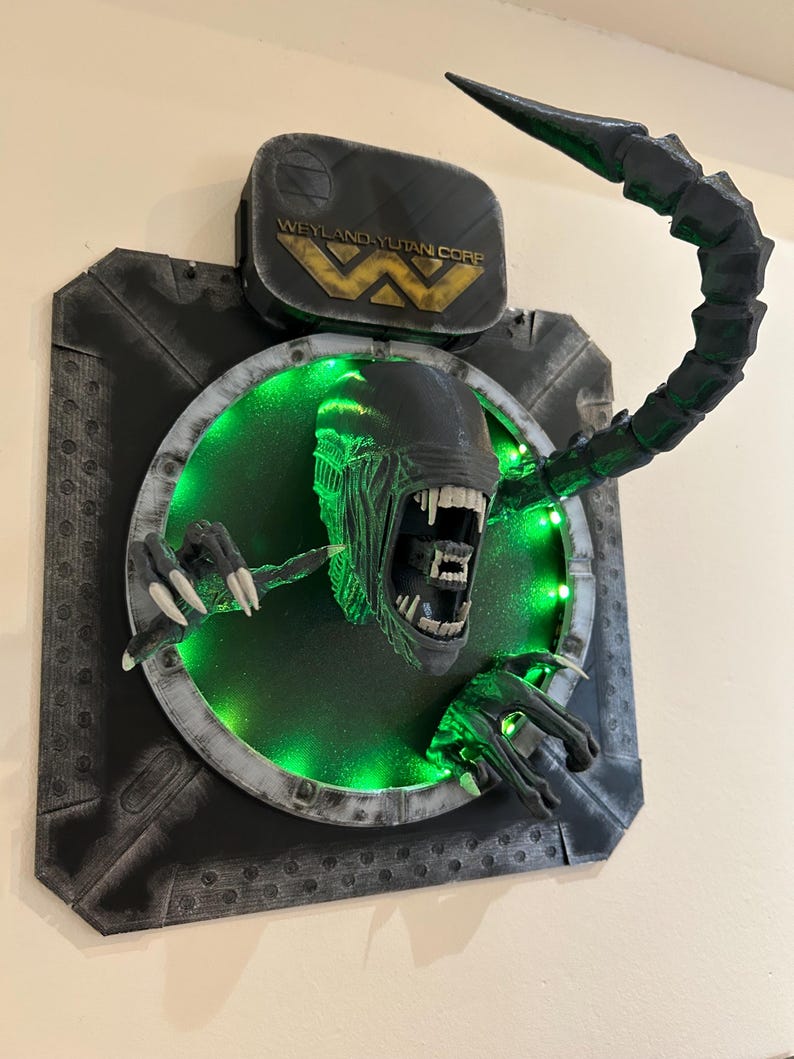 May include: A black and green 3D printed wall sculpture of an alien creature bursting through a circular opening. The alien has a long, segmented tail and sharp claws. The opening is lit with green LED lights and features a Weyland-Yutan Corp. logo above it.