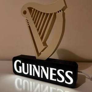 May include: A black and white illuminated sign with the word "GUINNESS" in white letters. The sign features a 3D harp in a cream colour.