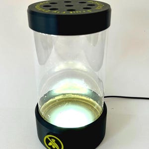 Może przedstawiać: Cylindryczna pułapka na muszki owocówki z przezroczystym plastikowym korpusem, czarną podstawą i czarną pokrywą z okrągłymi otworami wentylacyjnymi. Na pokrywie znajduje się napis BOFATECH FRUIT FLY EXTERMINATOR oraz żółta ikona.