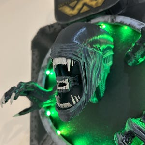 May include: A 3D printed model of an alien creature from the movie Alien, with a green glowing background and the Weyland-Yutani Corp logo. The alien is shown with its mouth open, revealing sharp teeth.