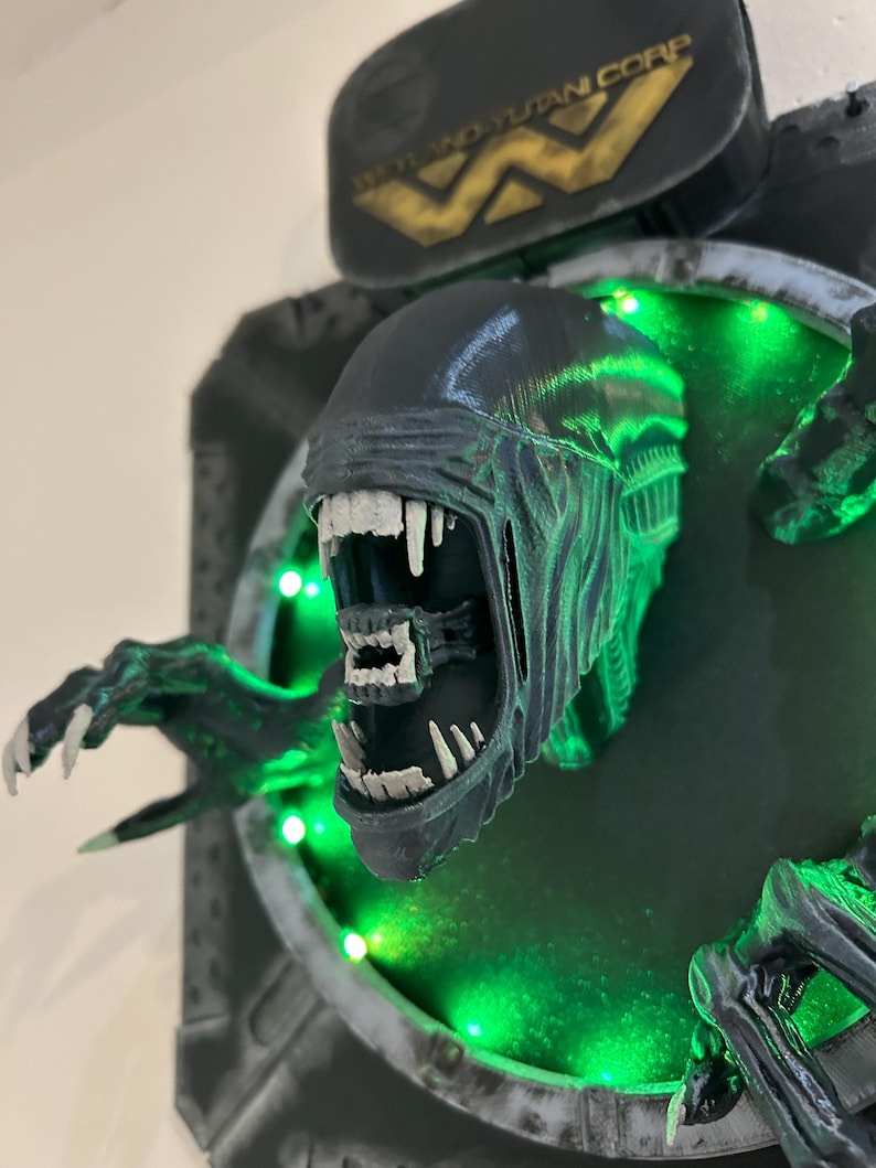 May include: A 3D printed model of an alien creature from the movie Alien, with a green glowing background and the Weyland-Yutani Corp logo. The alien is shown with its mouth open, revealing sharp teeth.