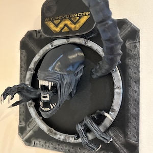 May include: A black and gray 3D printed wall sculpture of an alien creature from the movie Alien. The alien is bursting through a circular metal frame with the Weyland-Yutani Corp logo above it.