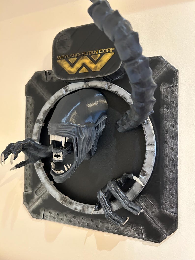 May include: A black and gray 3D printed wall sculpture of an alien creature from the movie Alien. The alien is bursting through a circular metal frame with the Weyland-Yutani Corp logo above it.