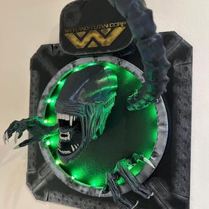 May include: A 3D printed wall sculpture of the Xenomorph from the Alien franchise. The sculpture is painted black and green and features a glowing green light behind the creature. The sculpture is mounted on a black and gray wall plaque with the Weyland-Yutani Corp logo.