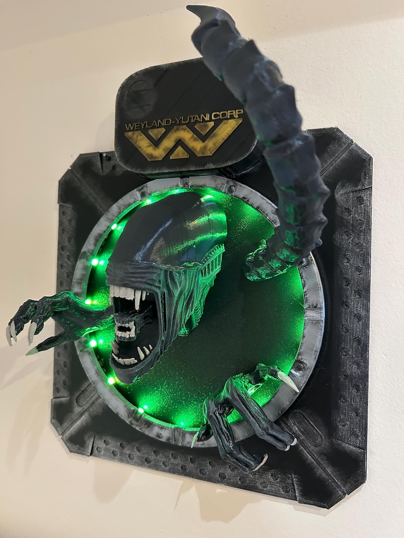 May include: A 3D printed wall sculpture of the Xenomorph from the Alien franchise. The sculpture is painted black and green and features a glowing green light behind the creature. The sculpture is mounted on a black and gray wall plaque with the Weyland-Yutani Corp logo.