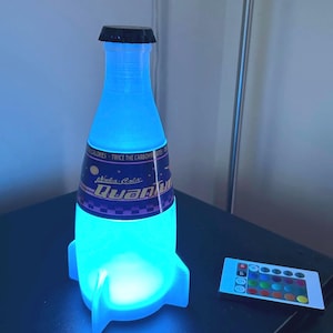 May include: A blue and white rocket-shaped lamp with a Nuka-Cola Quantum label. The lamp is lit up in a bright blue color and has a remote control with multiple color options.