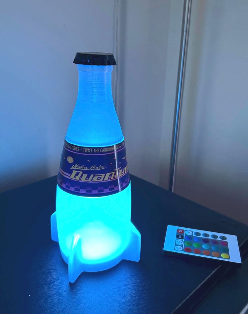 May include: A blue and white rocket-shaped lamp with a Nuka-Cola Quantum label. The lamp is lit up in a bright blue color and has a remote control with multiple color options.