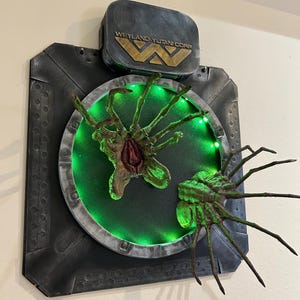 Peut inclure: Une œuvre d'art murale détaillée représentant une créature extraterrestre verte et marron avec de multiples membres, sur un fond vert éclairé. L'œuvre comprend le texte "WEYLAND-YUTANI CORP" en lettres dorées, dans un cadre gris foncé.