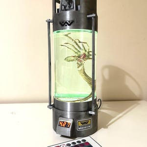 Peut inclure: Un récipient argenté et noir avec un cylindre en verre transparent contenant un liquide vert et une créature à plusieurs pattes. Le récipient comporte un affichage numérique indiquant le nombre 823 et un logo Weyland-Yutani. Une télécommande avec plusieurs boutons colorés est au premier plan.